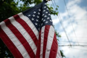 Picture of the flag of the United States at an angle. The background is a blue sky dotted with clouds and a tree. The flag is flapping.